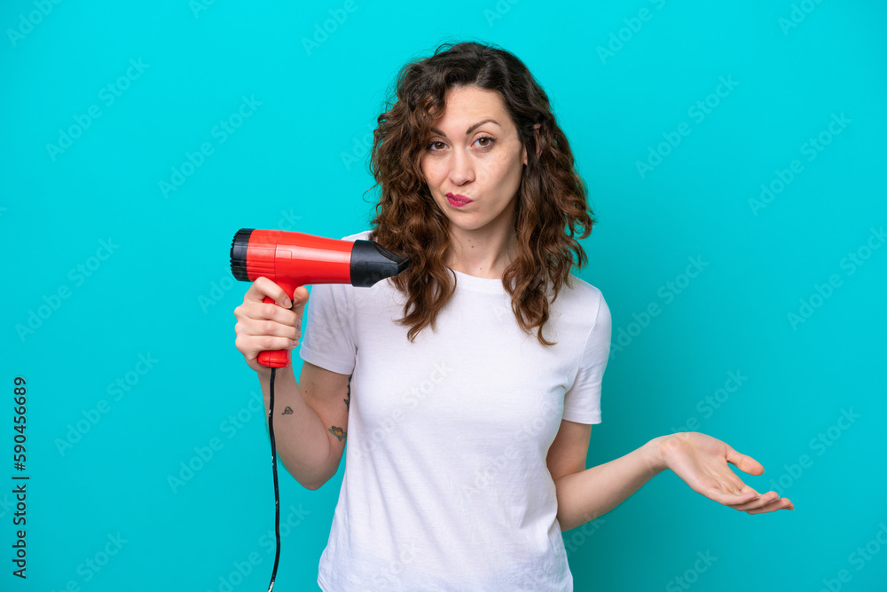 Young caucasian woman holding a hairdryer isolated on blue background making doubts gesture while lifting the shoulders Young caucasian woman holding a hairdryer isolated on blue background making doubts gesture while lifting the shoulders