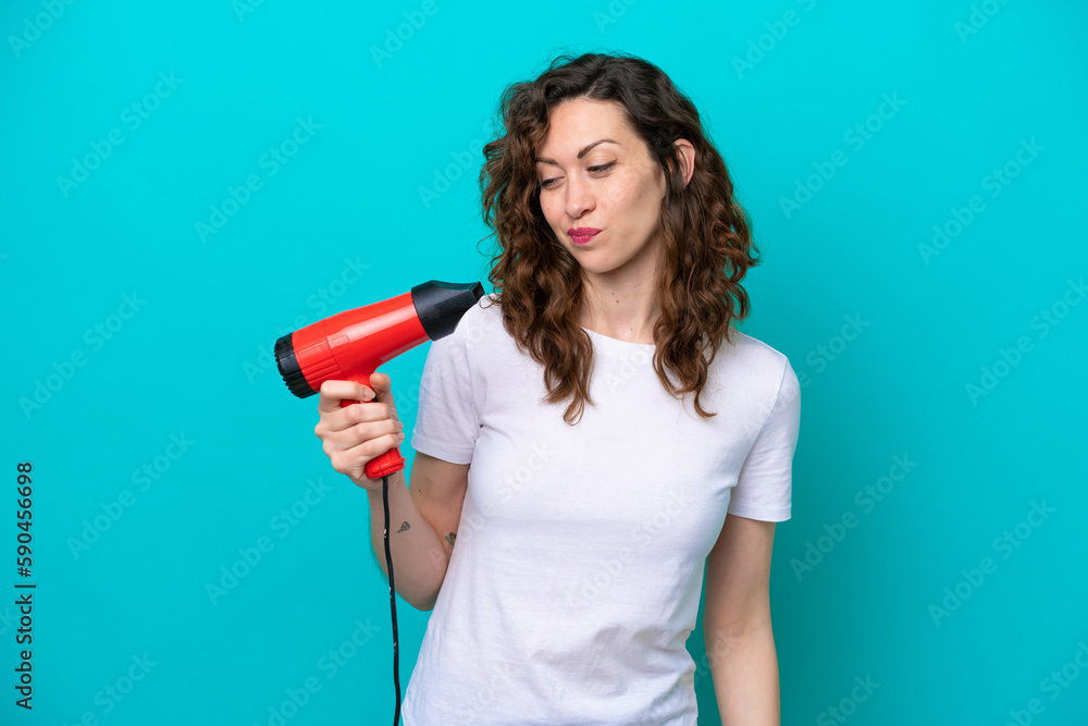 Young caucasian woman holding a hairdryer isolated on blue background with sad expression Young caucasian woman holding a hairdryer isolated on blue background with sad expression