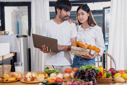 Young Asian couple cooking with fruits and vegetables and using laptop in the kitchen To cook food together within the family happily, family concept.