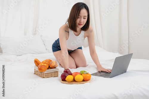 Portrait of white Asian woman with a tattoo sitting on the bed relaxing and playing laptop on weekend There was bread and fruit and red apples on a white bed to eat. holiday concept