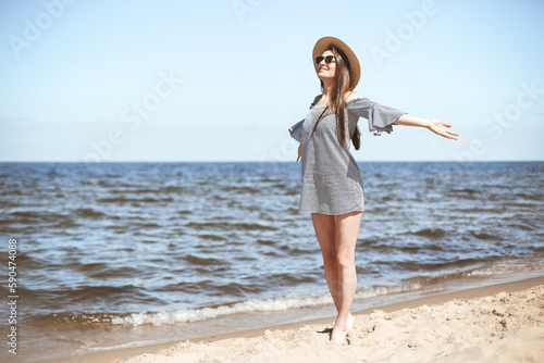Happy smiling woman in free bliss on ocean beach standing with open hands. Brunette female model in sunglasses and hat enjoying nature during travel holidays vacation outdoors, medium shot