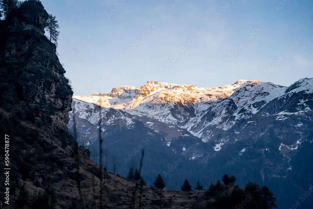 Sunlight lit peaks covered with snow in the distance with tree covered mountains in the foreground in manali