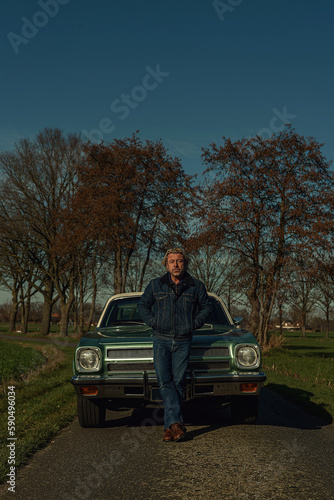 Man stands in front of a vintage american muscle car in sunny countryside. © ysbrandcosijn