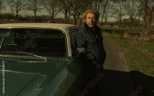 Man with blond hair in jeans stands on drivers side of a vintage american muscle car in sunny countryside.
