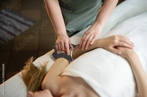 Hands of the masseur are close-up using a metal massage tool to massage the body of a woman lying on a couch. Health concept, body care, skin care, wellness