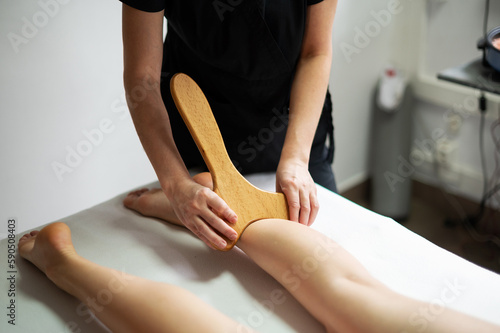 Hands of the masseur are close-up using a wooden massage tool to massage the leg of a woman lying on a couch. Health concept, body care, skin care, wellness