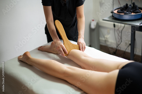 Hands of the masseur are close-up using a wooden massage tool to massage the leg of a woman lying on a couch. Health concept, body care, skin care, wellness