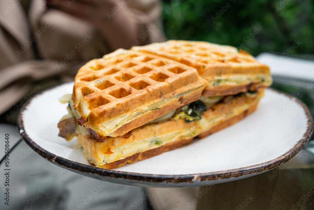 Waffle sandwich with spinach, mushrooms and cheese in a plate outdoor in sidewalk cafe. Close up. Soft focus
