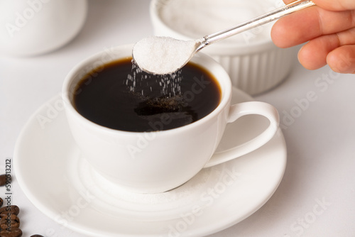 The process of pouring white sugar from a spoon into a white cup of coffee. Sugar addict, diabetes.