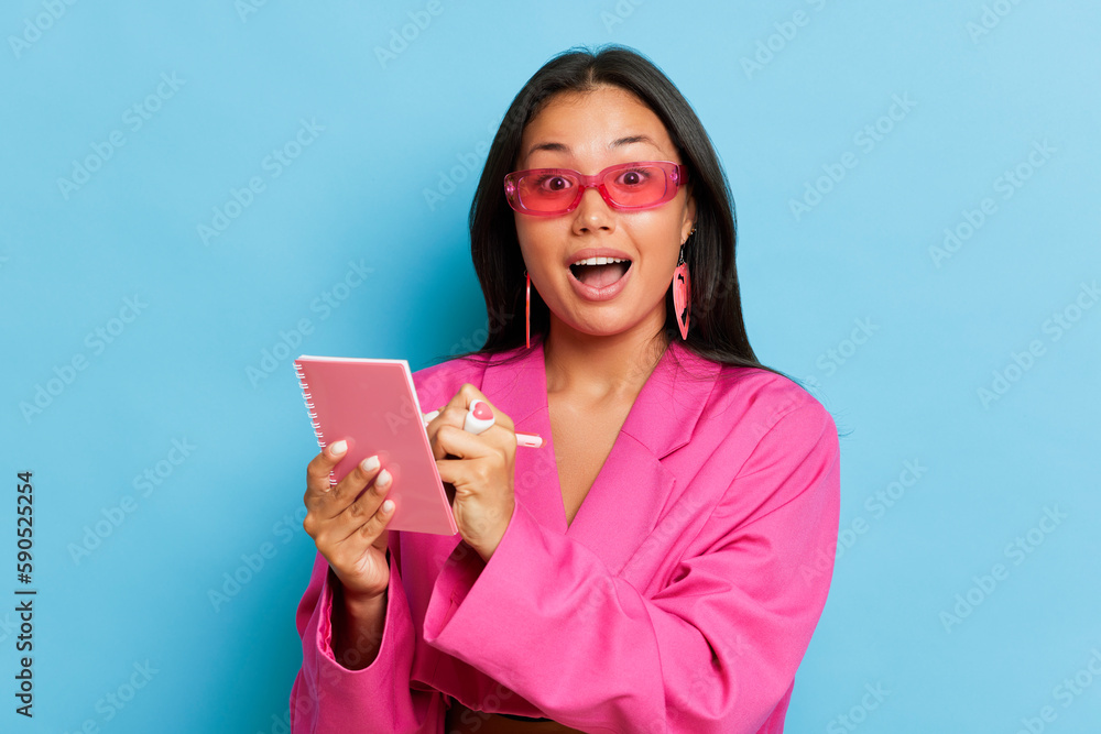Young happy woman in pink jacket looks at camera smiling, she holds notebook and a pen recording something, write down coordinates of desired company, copy space, business concept, high quality photo Young happy woman in pink jacket looks at camera smiling, she holds notebook and a pen recording something, write down coordinates of desired company, copy space, business concept, high quality photo