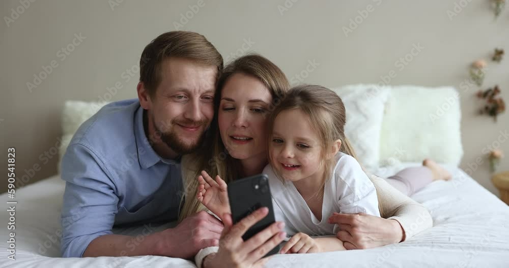 Cheerful cute daughter kid and happy couple of young parents resting on bed, taking selfie photo on smartphone, watching, shooting video for social media, making video call Cheerful cute daughter kid and happy couple of young parents resting on bed, taking selfie photo on smartphone, watching, shooting video for social media, making video call