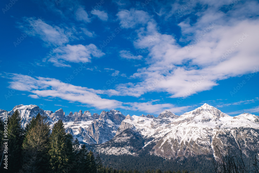 A mountain range with snow capped mountains in the background and a blue sky with clouds above it and a few trees in the foreground, Dolomiti, Italy A mountain range with snow capped mountains in the background and a blue sky with clouds above it and a few trees in the foreground, Dolomiti, Italy