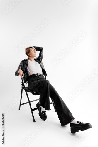 Full body of smiling young female in trendy black pants and leather jacket touching blond hair and relaxing on chair against white background