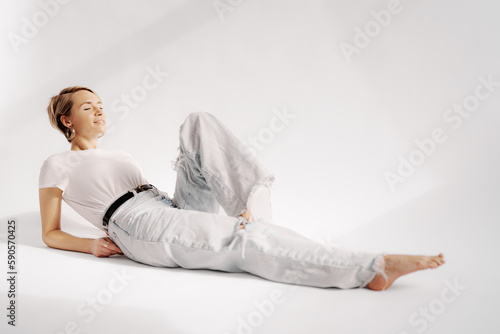 Side view of young female lying on floor with raised leg and back while using support of hands and looking away against white background