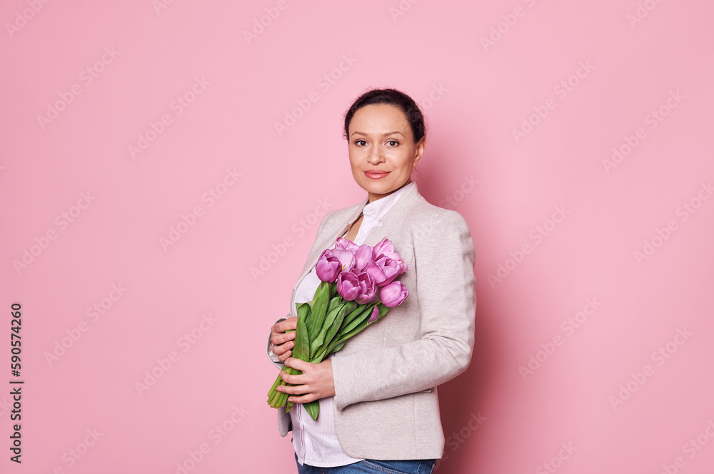 Portrait of a beautiful pregnant middle-aged woman in casual clothes, holding a bouquet of purple tulips, smiling at camera, isolated on pink background. Happy Mother's Day. International Women's Day Portrait of a beautiful pregnant middle-aged woman in casual clothes, holding a bouquet of purple tulips, smiling at camera, isolated on pink background. Happy Mother's Day. International Women's Day