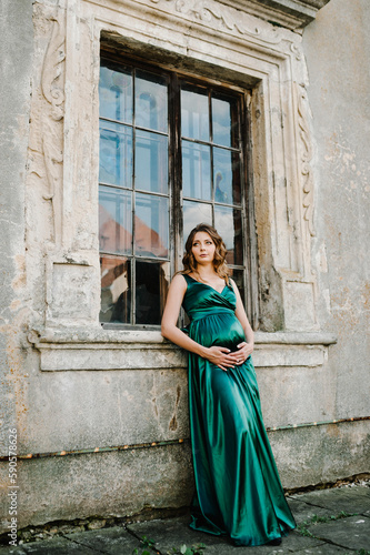 Pregnant woman wearing long green dress, holding tummy near old window. New life concept.