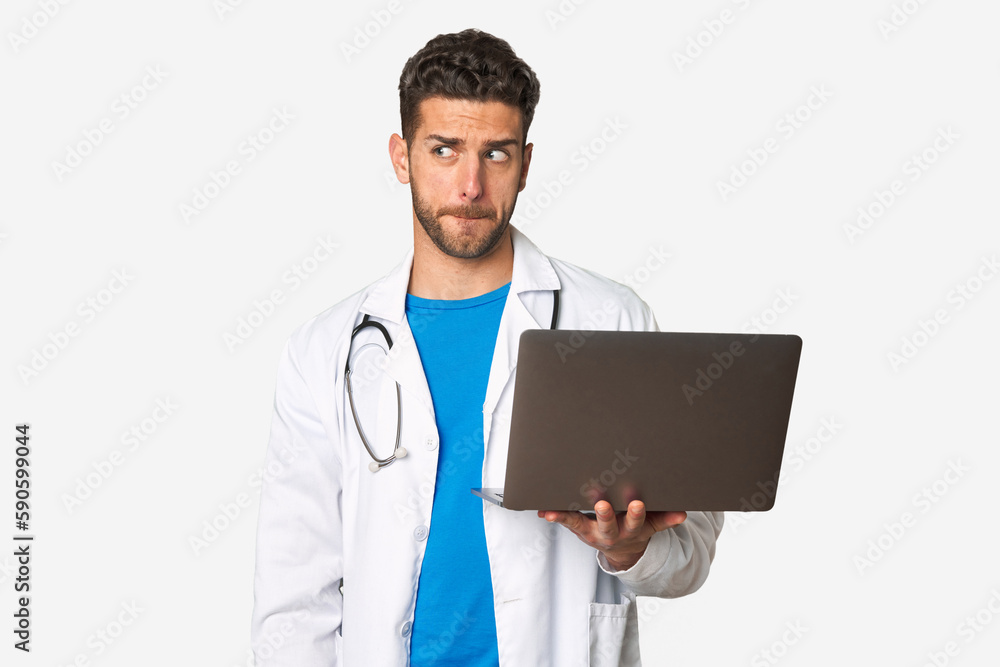 Young doctor man holding a laptop confused, feels doubtful and unsure. Young doctor man holding a laptop confused, feels doubtful and unsure.