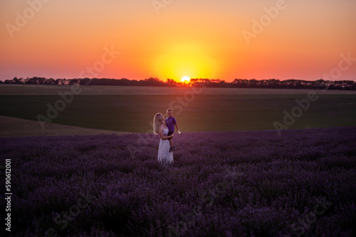 Millenial young mother holds little son in arms at sunset in purple lavender field. Walks in the countryside. Trust, protection of the child. Happy childhood. Allergy concept, single parent. Nature