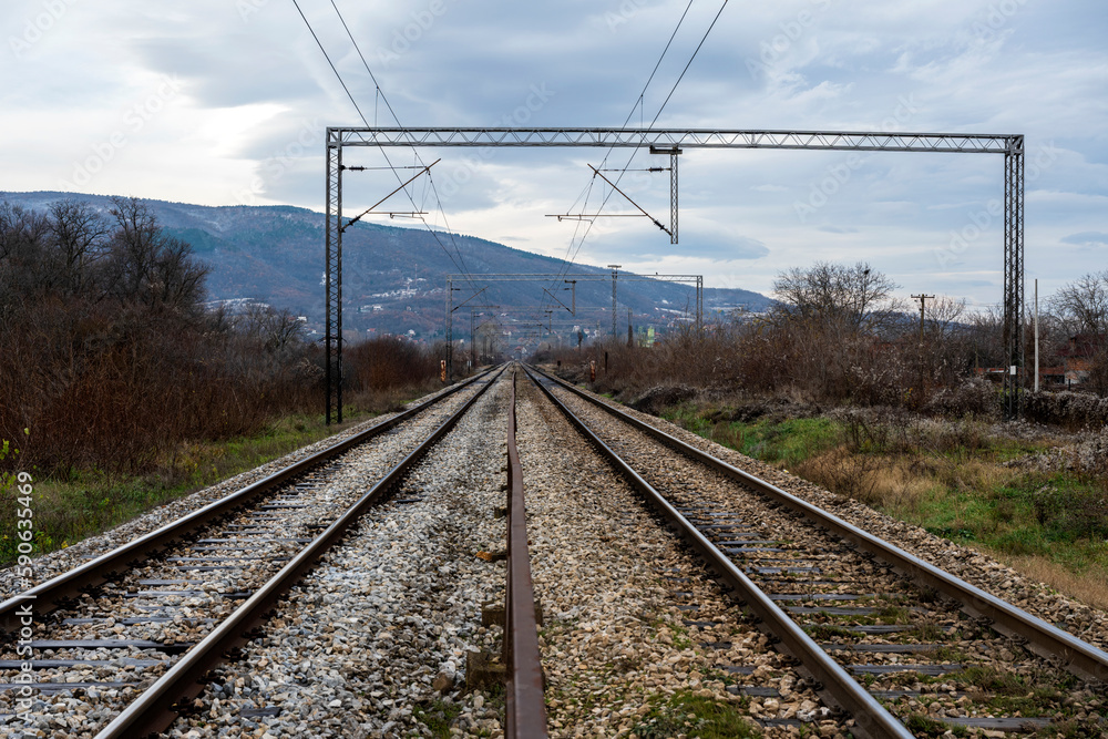 Infrastructure development project with laying of railway tracks equipped with overhead electric poles in parallel to high powered supply lines in dry terrain during summertime