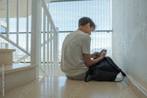 Sad teenage boy with mobile phone and backpack sitting on stairs. Teenager surfing on Internet, watching video, using app. Education, learning difficulties, mobile addiction concept