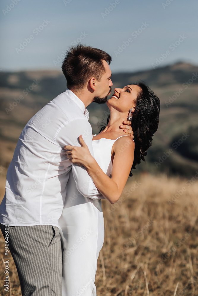 A stylish model couple in the mountains in the summer. A young boy and a girl in a white silk dress are walking on the slope against the background of the forest and mountain peaks. boy kisses a girl A stylish model couple in the mountains in the summer. A young boy and a girl in a white silk dress are walking on the slope against the background of the forest and mountain peaks. boy kisses a girl