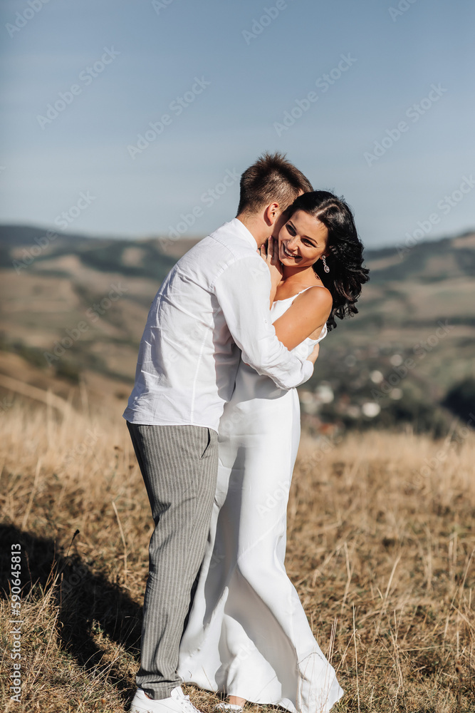 A stylish model couple in the mountains in the summer. A young boy and a girl in a white silk dress are walking on the slope against the background of the forest and mountain peaks. boy kisses a girl