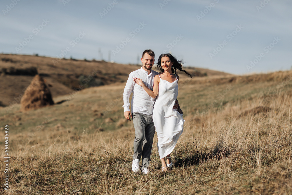 A stylish model couple in the mountains in the summer. A young boy and a girl in a white silk dress are walking on the slope against the background of the forest and mountain peaks. boy kisses a girl A stylish model couple in the mountains in the summer. A young boy and a girl in a white silk dress are walking on the slope against the background of the forest and mountain peaks. boy kisses a girl