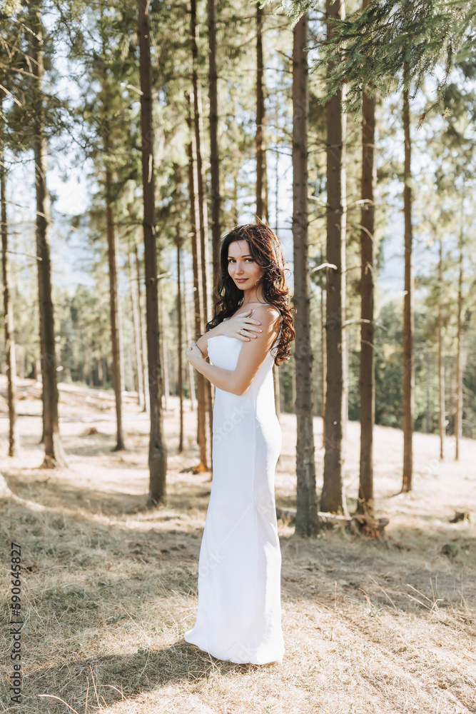 A girl with long black curly hair blowing in the wind is standing in a white silk dress. Standing in the spring forest against the background of large trees