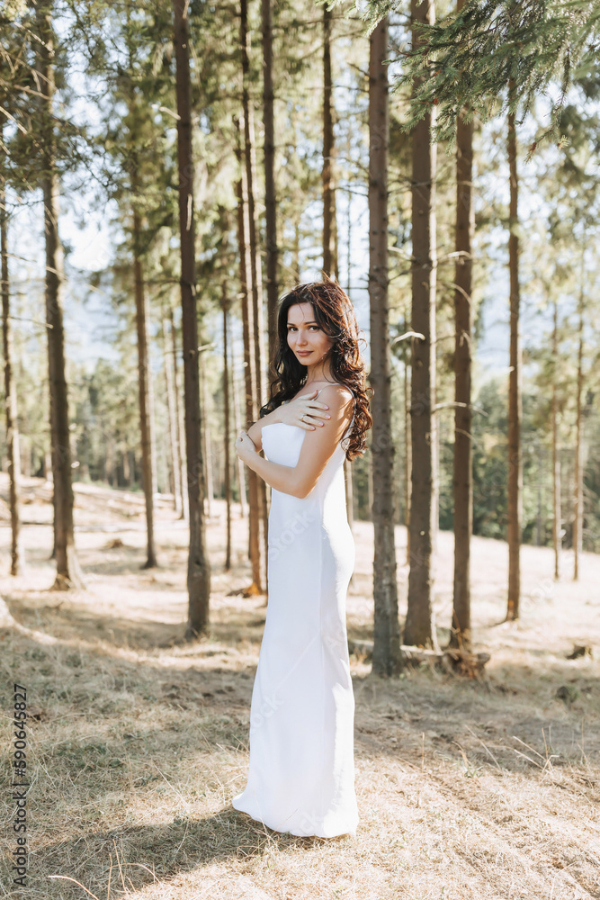 A girl with long black curly hair blowing in the wind is standing in a white silk dress. Standing in the spring forest against the background of large trees A girl with long black curly hair blowing in the wind is standing in a white silk dress. Standing in the spring forest against the background of large trees