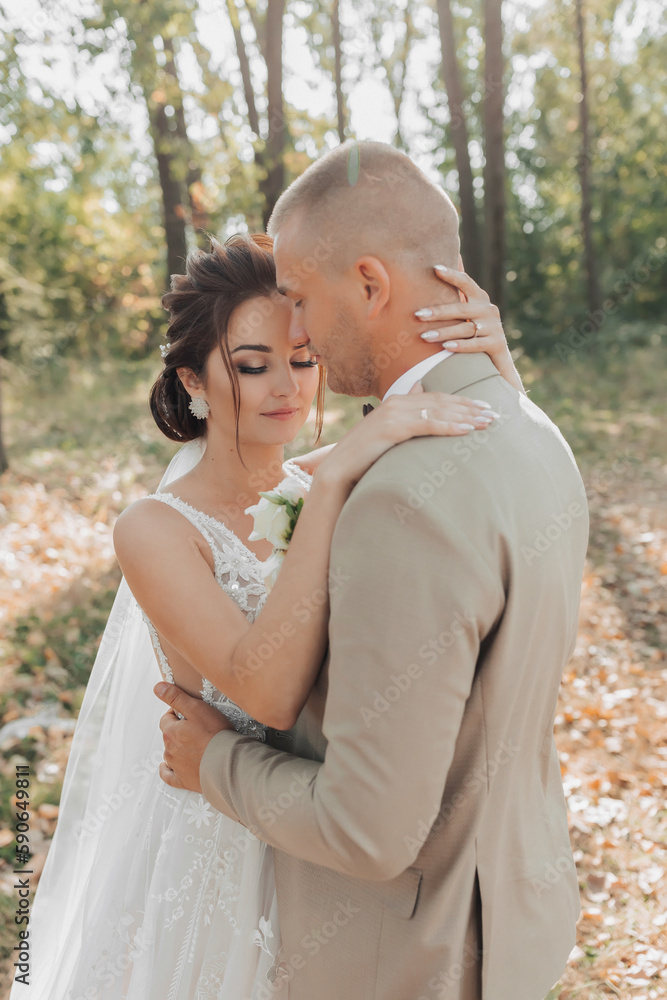 Wedding portrait photo. Groom in a light suit. The bride and groom are standing in the forest embracing. Beautiful makeup and hair. Couple in love. Summer light. The shadows