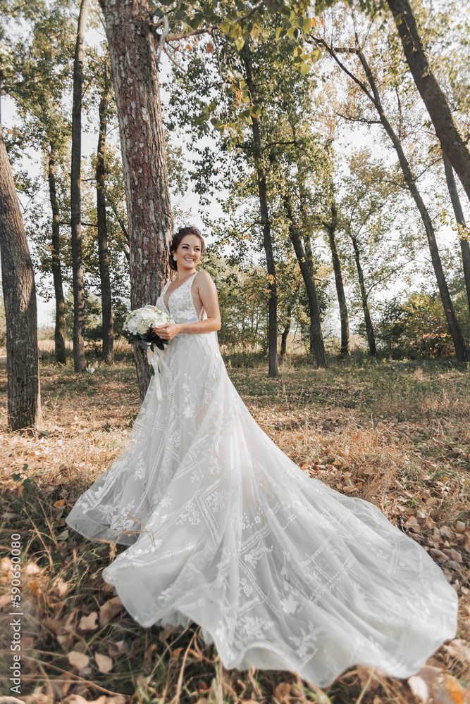 Wedding photo in nature. The bride is standing in the forest. The bride in a beautiful dress with a long train, holding her bouquet of white roses, smiling sincerely at the camera. Portrait
