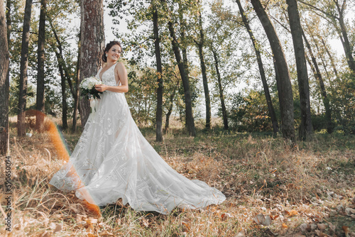 Wedding photo in nature. The bride is standing in the forest. The bride in a beautiful dress with a long train, holding her bouquet of white roses, smiling sincerely at the camera. Portrait