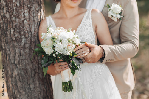 Photo of a wedding bouquet of white flowers and greenery in the hands of the bride and groom. The groom holds the bride's hand. Beautiful hands. French manicure. Wedding rings