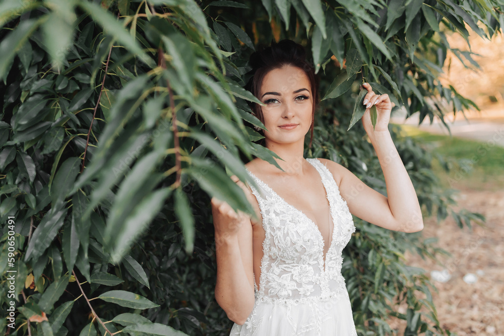 Wedding photo on a green background. The bride in a white dress stands near green bushes, holds a leaf and looks into the lens. Beautiful makeup. Portrait of the bride Wedding photo on a green background. The bride in a white dress stands near green bushes, holds a leaf and looks into the lens. Beautiful makeup. Portrait of the bride
