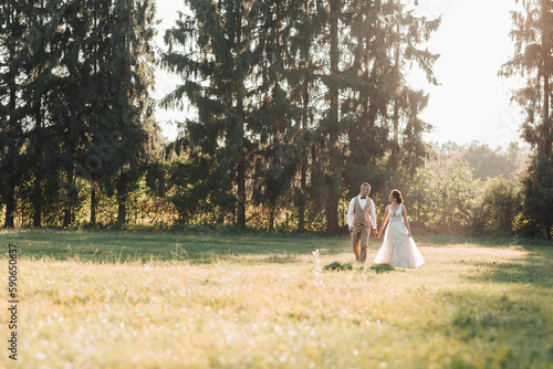Fototapeta Naklejka Na Ścianę i Meble -  Happy newlyweds are running in the field, holding hands, looking at each other, behind them are big green Christmas trees. Beautiful light. Stylish groom. Pretty Girl