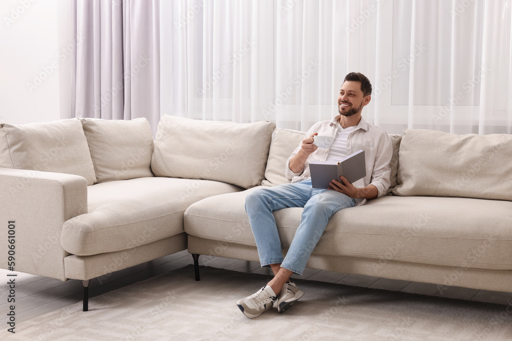 Happy man drinking coffee while reading book on sofa near window with beautiful curtains at home in living room. Space for text Happy man drinking coffee while reading book on sofa near window with beautiful curtains at home in living room. Space for text