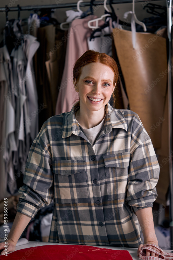 excited caucasian dressmaker woman sews clothes at tailor office, engaged in tailoring,redhead lady in checkered shirt posing at camera, fabric clothes in the background. dressmaking industry