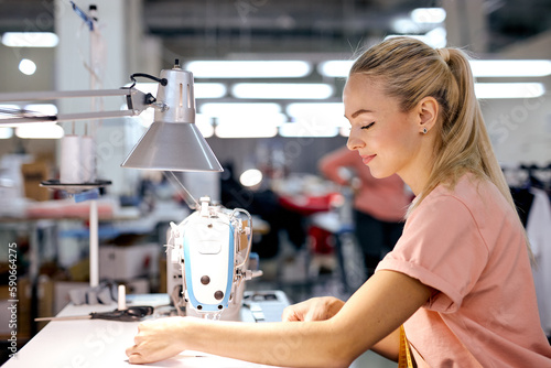 young nice caucaian blonde textile factory worker on production line using modern sewing machine, at workshop. hardworking woman engaged in sewing or tailoring, dressmaking concept photo