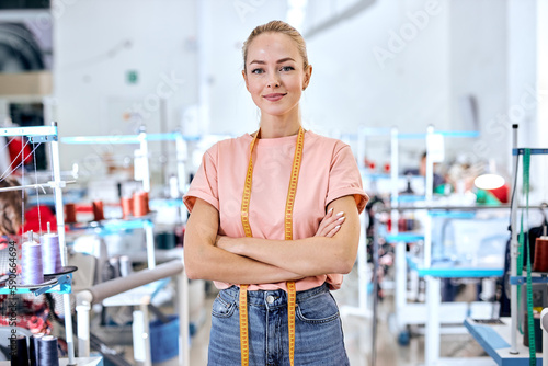 confident woman dressmaker standing in textile factory at workshop, attractive tailor after work stand posing, looking at camera smiling. female use measuring tape for sewing and making garments