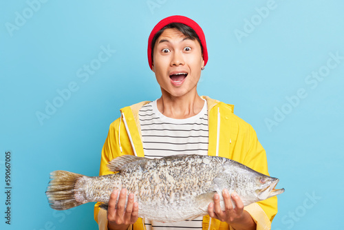Happy guy dressed in red cap and yellow jacket holding big fish in his hands, showing enjoyment by such a good catch, fortune fishing concept, copy space, high quality photo
