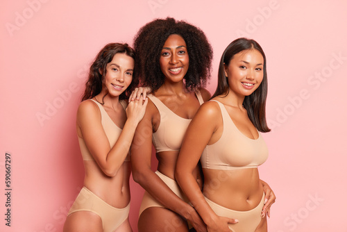 Three different women in lingerie stand half-turned in the studio with african american girl model in the middle, wearing a comfortable beige top and panties, underwear fashion concept, copy space