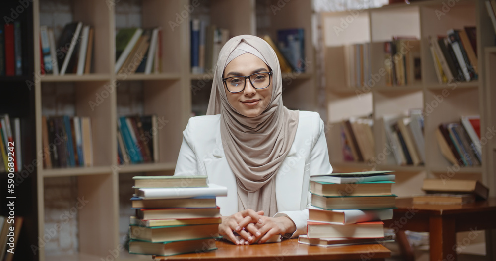Muslim student is doing research at desk full of books in library. Girl wearing hijab is preparing for exam or making a project, then looking at camera Muslim student is doing research at desk full of books in library. Girl wearing hijab is preparing for exam or making a project, then looking at camera