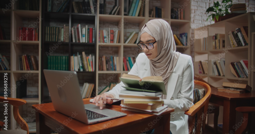 Female muslim student is studying at desk in library, using laptop and books. Girl wearing hijab is preparing for exams - student lifestyle, modern Islam concept 4k close Female muslim student is studying at desk in library, using laptop and books. Girl wearing hijab is preparing for exams - student lifestyle, modern Islam concept 4k close