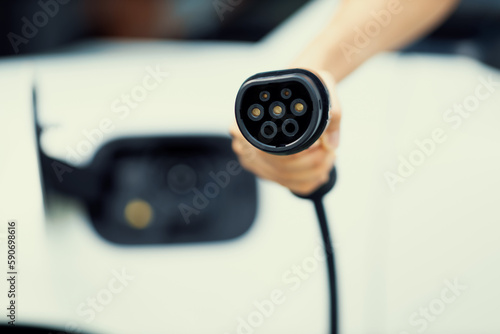 Progressive new alternative clean energy vehicle and electric charging device concept. Focus closeup hand holding and pointing EV charger plug at camear from electric charging station at camera photo