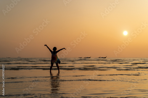 back asian child or kid girl standing on summer sea beach and happy raising hands with golden sunlight on evening sunset or morning sunrise with tourist fishing boat for vacation travel holiday relax