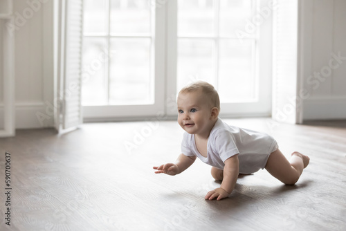 Happy baby in bodysuit crawling on knees on clean warm floor. Cheerful active little infant kid training physical skills, looking away, smiling. Pretty adorable infant kid home portrait