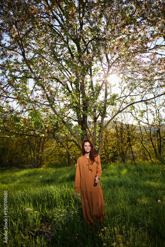 a sweet, modest woman with long hair stands in the countryside near a flowering tree and looks at the camera with her hands clasped together