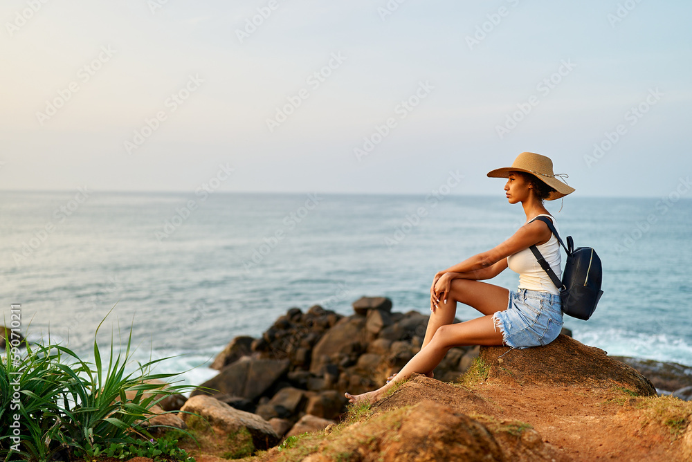 Multiethnic woman in straw hat enjoys tropical vacation sitting on rocky cliff with sea view. Black female with backpack sightseeing on scenic location. Pretty lady tanning on island on ocean sunrise.