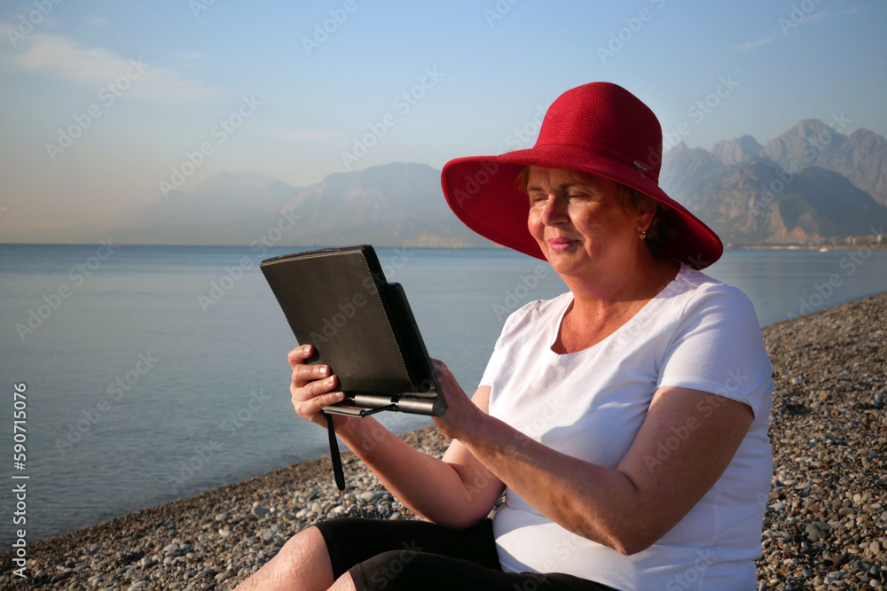 Senior woman holding tablet computer sitting on the beach, wearing in big red sunhat and taking sun bath