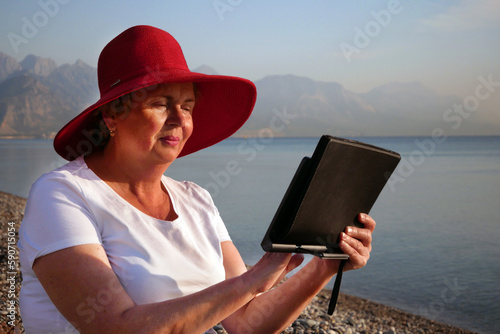 Senior woman holding tablet computer sitting on the beach, wearing in big red sunhat and taking sun bath © lesslemon